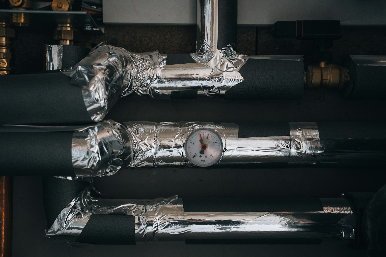 Insulated hot-water pipework and brass valves around a pressure gauge in a plant cupboard